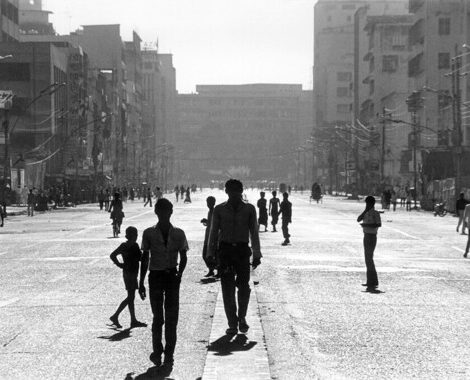 Silhouettes of people walking down wide open city street with buildings in the background.