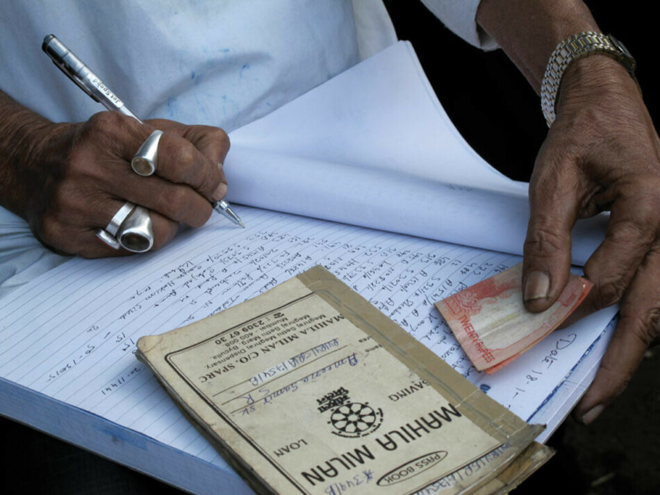 A person holding a pen in one hand, writing on white paper ledger while the other hand holds money next to a bank passbook. 