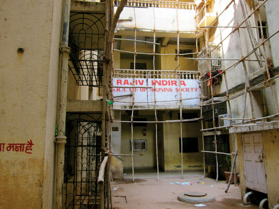 Urban setting, view down an alley with tall apartment-like buildings surrounded by scaffold with a white banner with red writing. 