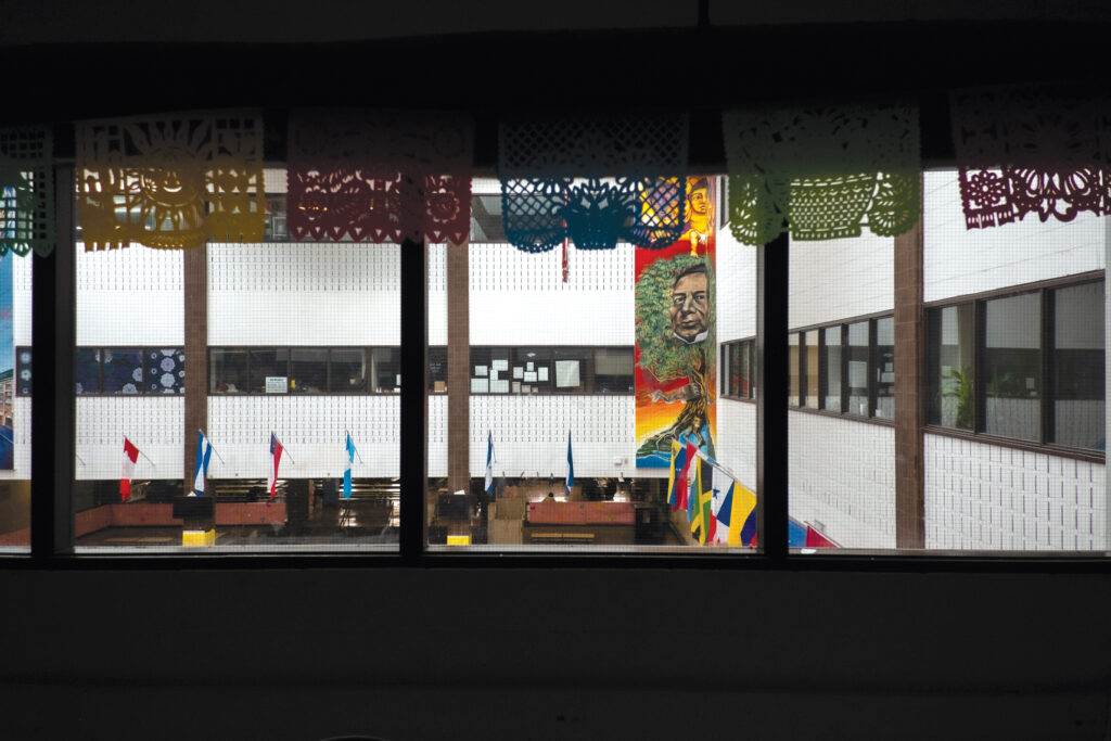 Photograph of windows that look out into the courtyard of a building. Courtyard is flaked with windows to other rooms in the building, a mural, flags, and cut out pieces of paper