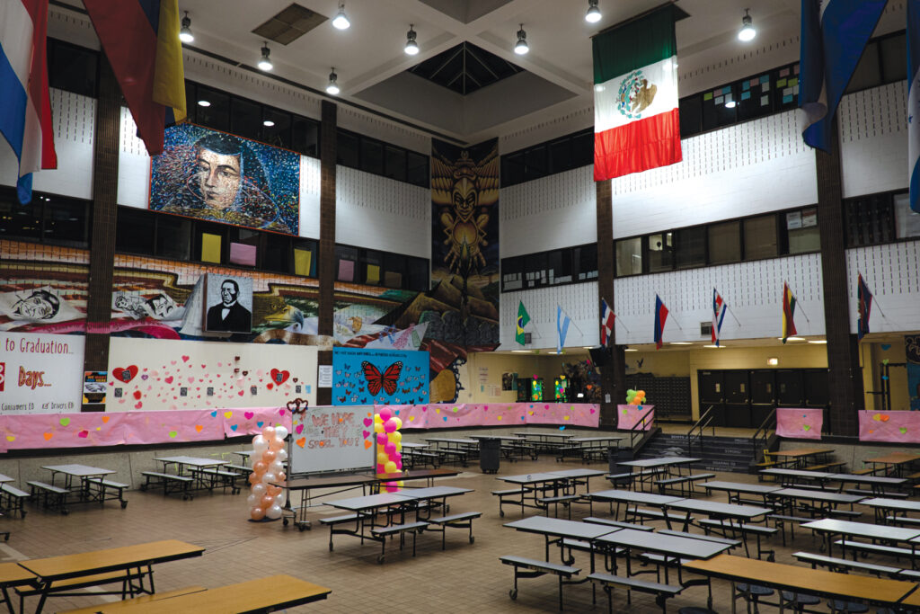 Interior of a cafeteria with high ceilings, tables and seats with murals on the walls and flags hanging from the ceiling