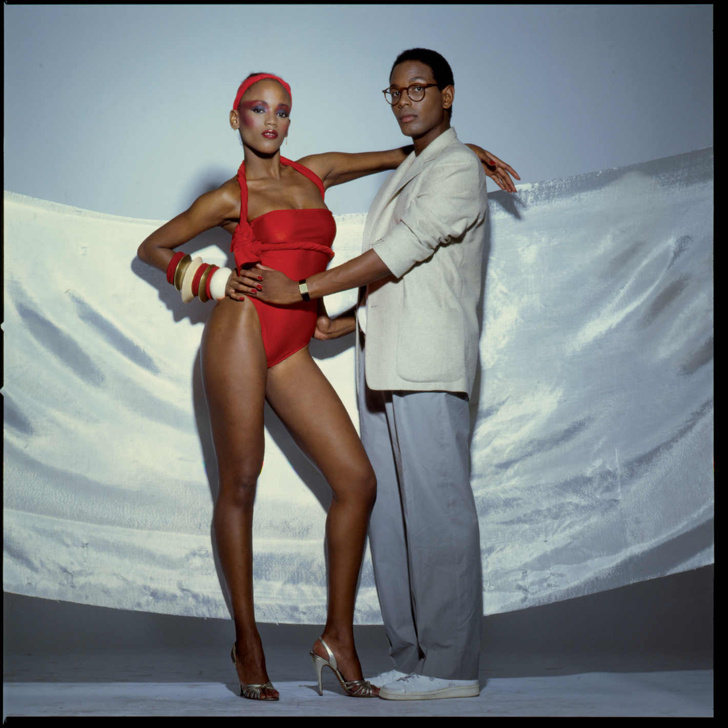 A man in glasses, a white jacket, in grey pants holds the torso of a woman dressed in a red bathing suit, wearing a number of bracelets on her wrist and a red bandana while in silver heels against a shimmery background