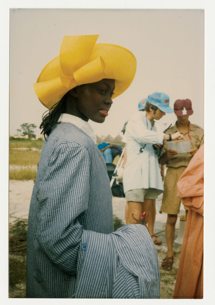 Photograph of a woman outside wearing a yellow hat and a jacket with blue and white stripes. Behind her are two other people in hats looking at a document