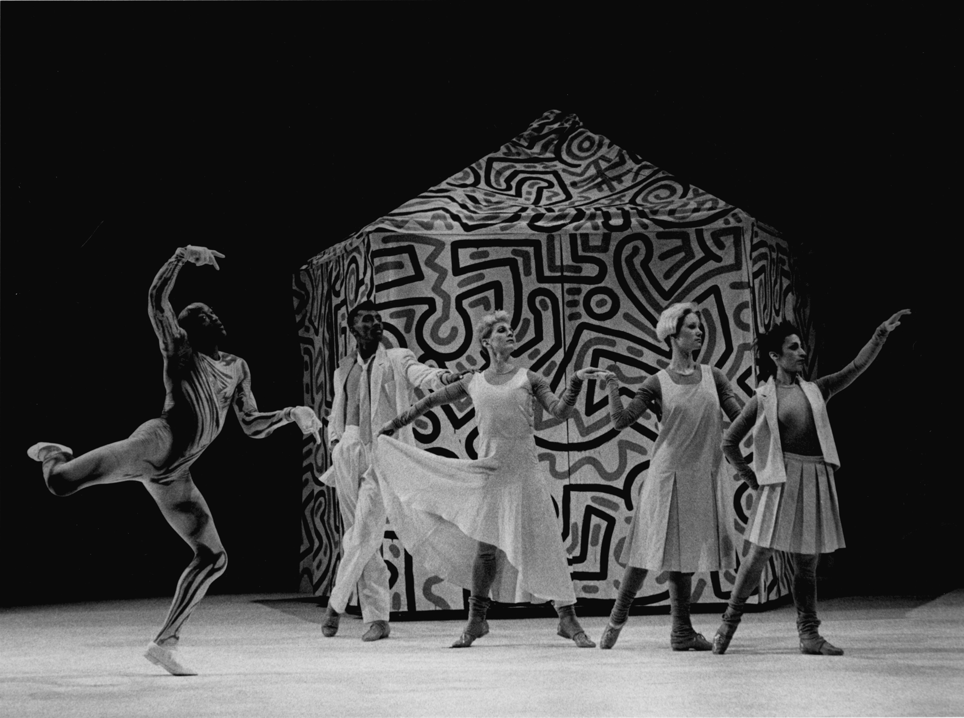 Black and white photograph of male and female dancers in white in front of a tent with squiggly lines all over it.