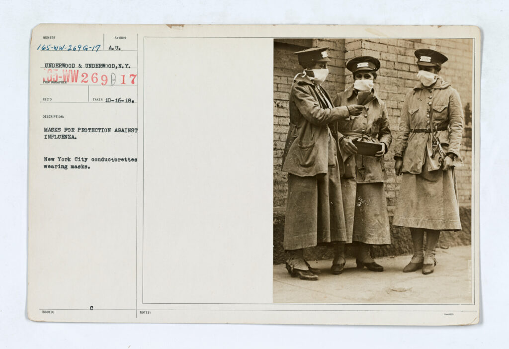 Card from a catalog with a vintage, photograph of three women wearing dress uniforms with jackets, masks, and hats talking with each other outside. Card reads a catalog number, underwood and underwood, masks for protection against influenza, New York City conductorettes wearing masks October 16, 1918