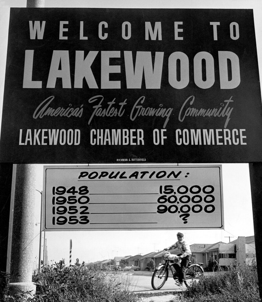 Black and white photograph of a sign that says welcome to lakewood, america's fastest growing community, lakewood chamber of commerce. A boy on a bike appears underneath with a view of homes in the background