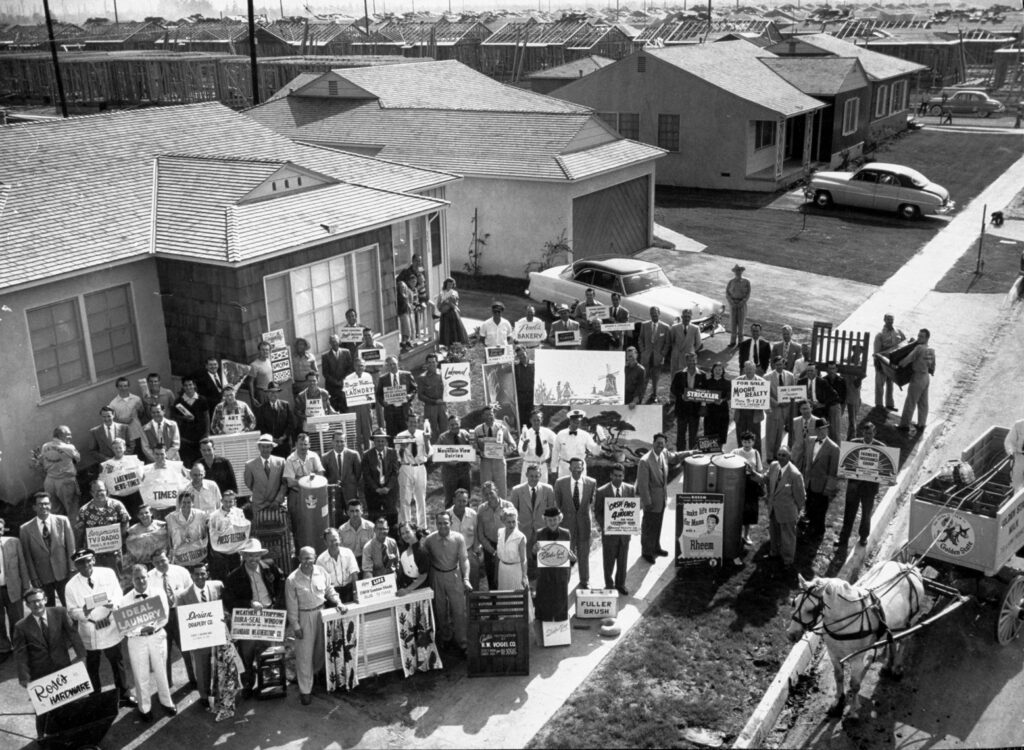 Black and white photograph of people standing in front of a home holding signs with a horse and buggy in front and two cars in the background