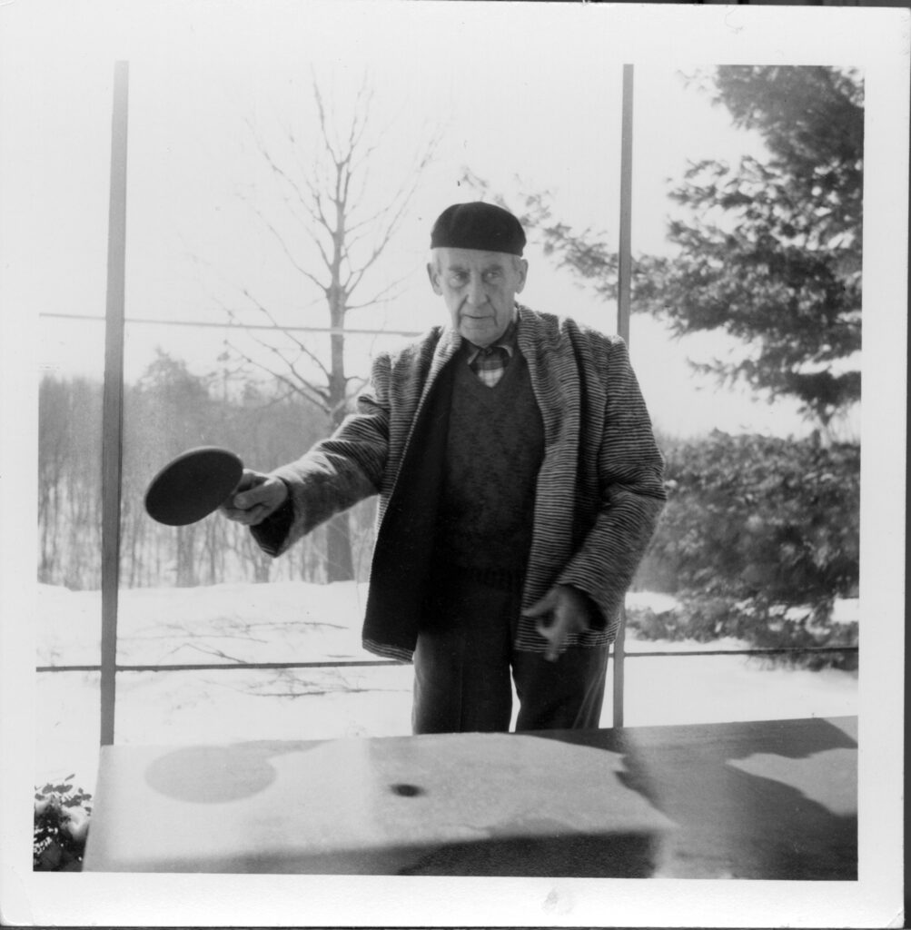 Black and white photograph of a man wearing a beret holding a racket in front of a table. A glass window behind him shows trees outside.