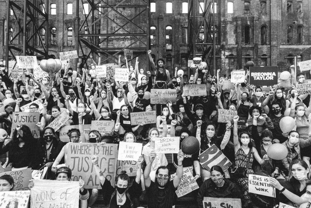 Black and white photograph of a crowd of people holding signs that say many things including where is the justice for all, Breonna Taylor, say her name, white silence to violence, this is essential work