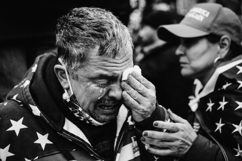 Black and white photograph of an older man wearing a sweatshirt with white stars, holding a cloth to his eyes as he sheds white tears while the woman next to him holds his arm