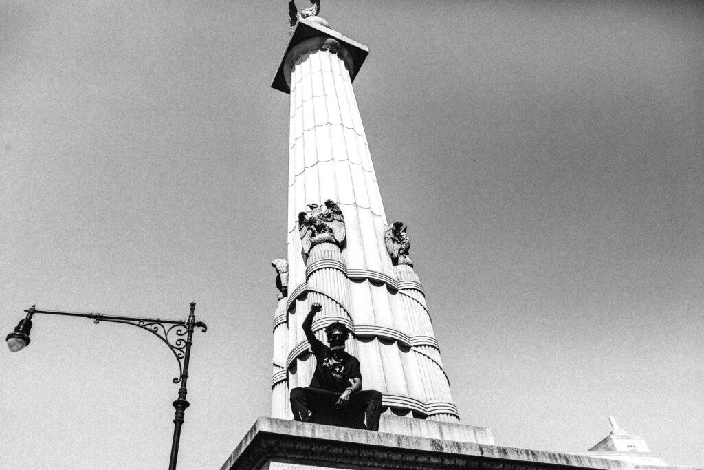Black and white photograph of a pilar like sculpture with a man sitting at the base with a mask on and his fist in the air
