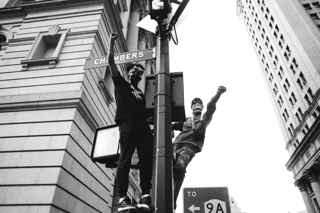 Black and white photograph of two people wearing masks standing on the edge of a lamp post with a street sign that says Chambers Street with a concrete building to the left and a skyscraper to the right with a sign to route 9A behind them