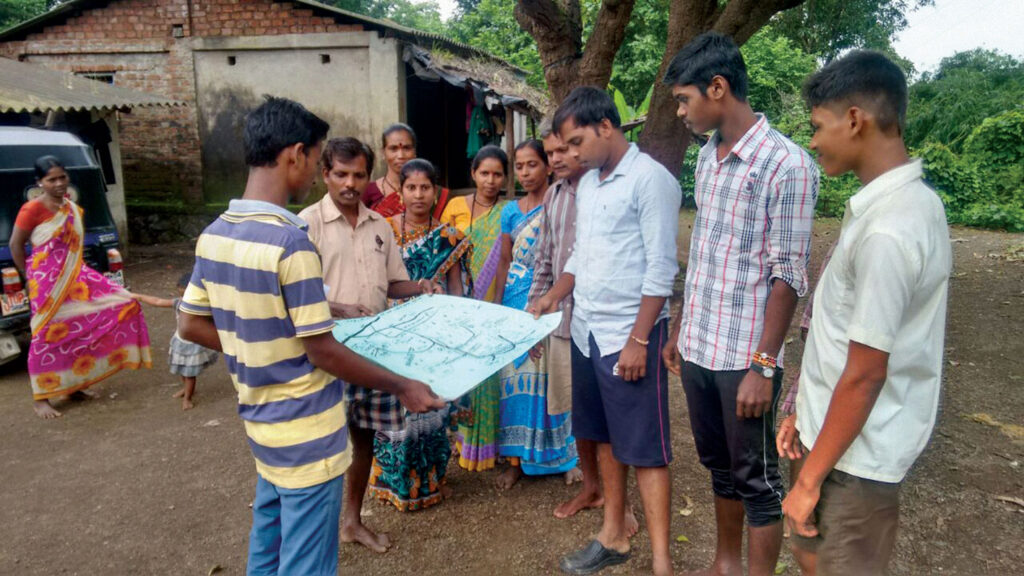 Men and women in an outdoor setting under a tree look at a white map a man is holding. 