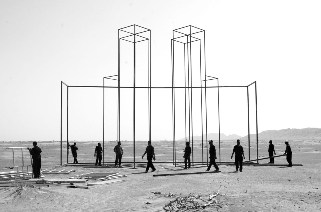 Black and white photograph of men in a desert building a tall scaffold inthe shape of a building.