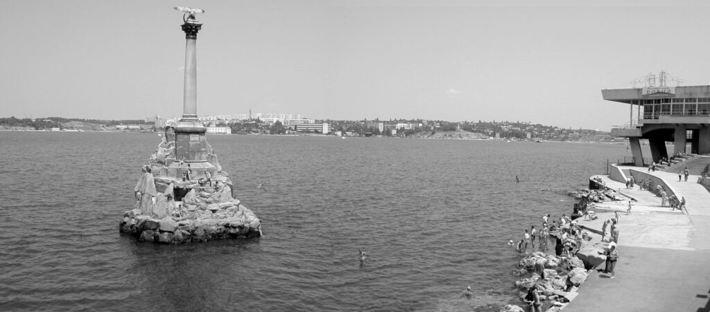 Daytime view of a recreational area above water with people jumping into the water across from a statue on rocks in the water