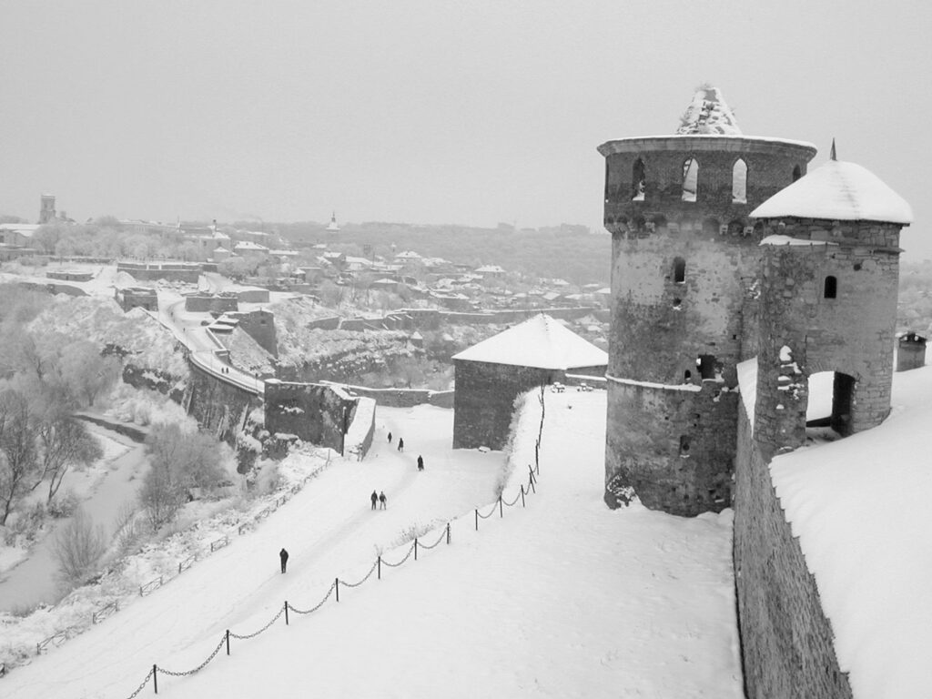 Daytime photograph of a hollowed out stone structure covered with and surrounded by snow. People below walking a snow-cover trail toward another part of the mountain.