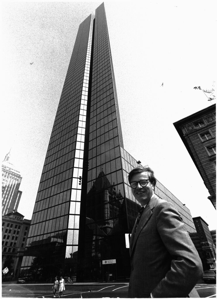 Man in glasses and a suit standing in front of a very tall skyscraper