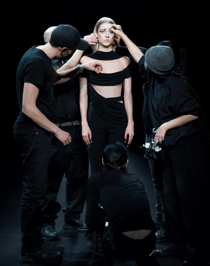 A model in black being surrounded by three people wearing black preparing for a show against a dark background