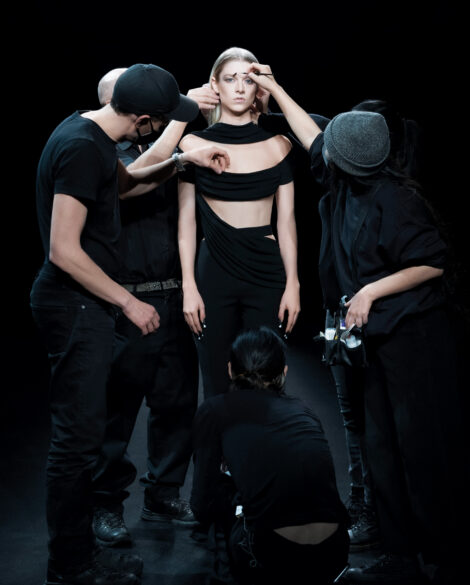 photograph of a model standing wearing a black outfit with blonde hair surrounded by three other people tending to attire and makup