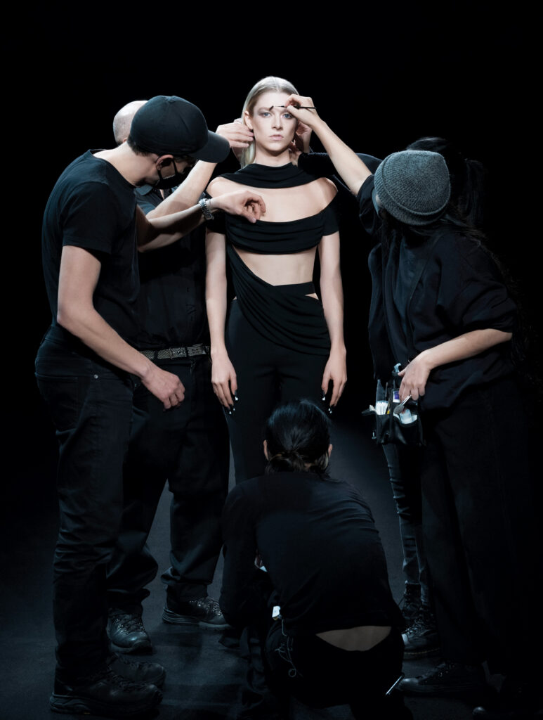 photograph of a model standing wearing a black outfit with blonde hair surrounded by three other people tending to attire and makup