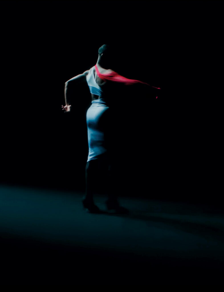 A figure in blue and red fabric dancing against a dark background