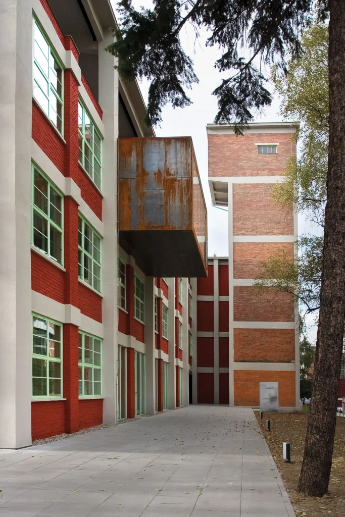 Vertical photograph of a three story brick building with a grid of white painted brick above green window frames with a rusted metal rectangle object extending out over a paved walkway with trees to the right.