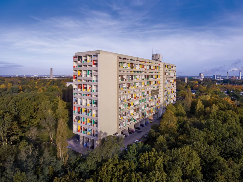 Aerial view of a tall, rectangular concrete apartment building with colorful balconies surrounded by a lush landscape of trees on a sunny day surrounded by blue sky