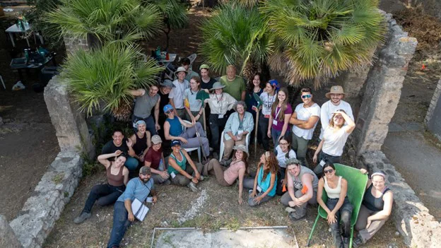 Aerial group photo of 25 people with Roman ruins and palm trees.