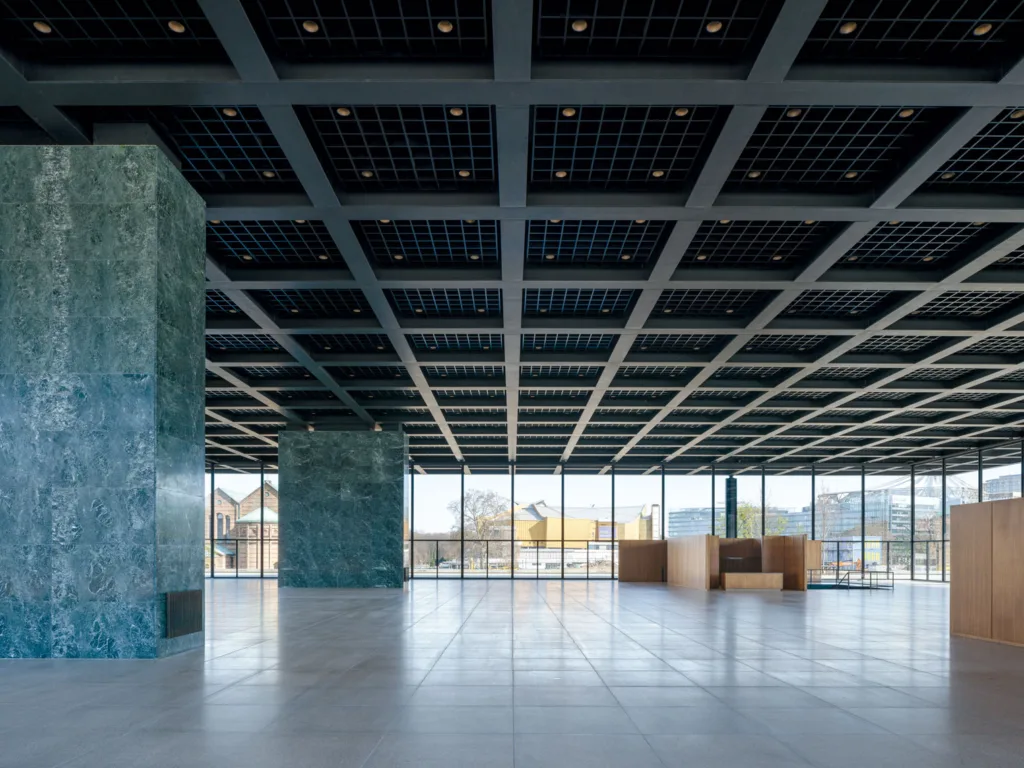 Color photograph of the interior of a spacious empty gallery surrounded by windows with a grid structure on the ceiling and thick square marble columns on the left.