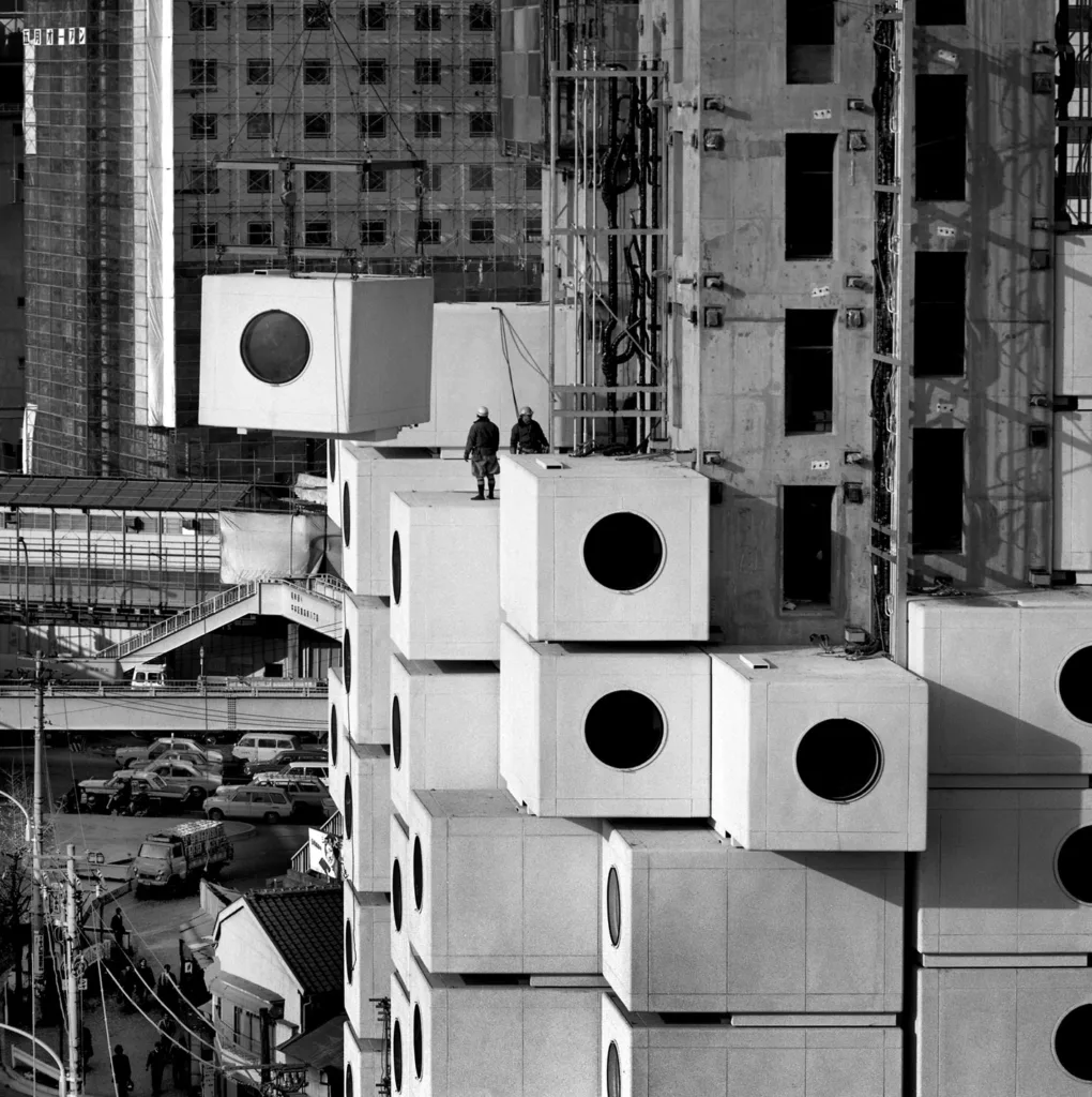 Black and white photograph of large concrete square structures with circle windows being lifted by crane and stacked on similar concrete blocks with two people in hard hats observing.