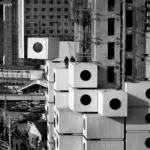 Black and white photograph of large concrete square structures with circle windows being lifted by crane and stacked on similar concrete blocks with two people in hard hats observing.