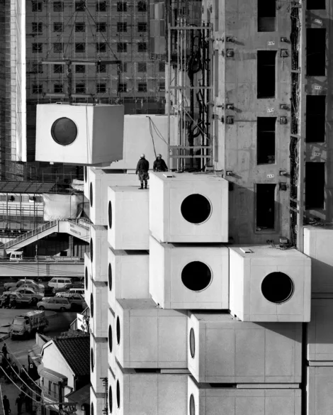 Black and white photograph of large concrete square structures with circle windows being lifted by crane and stacked on similar concrete blocks with two people in hard hats observing.
