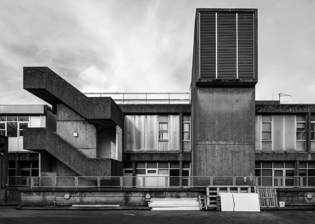 Black and white photograph of a 2 story concrete building with diagonal stairs on the left and a tall tower with a large vent on the right with palelts and wood stacked in the foreground.