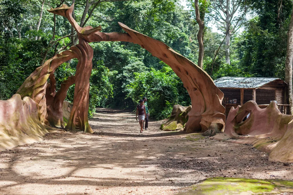 Two people walk beneath a large wooden arch created by a decoratively carved tree trunk. 