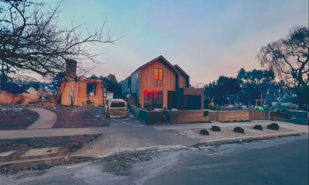 Photograph of a wooden clad home with windows that reflect the sunset in the distance surrounded by homes damaged by fire.