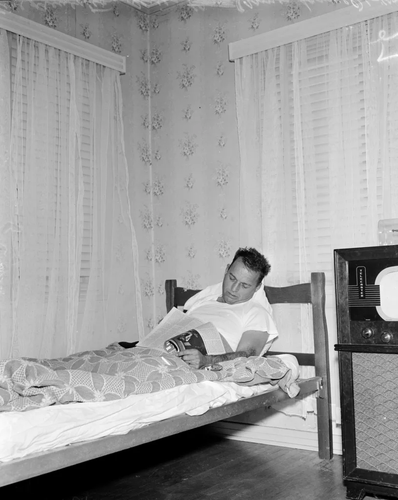 Black and white photograph of a man sleeping in a twin bed covered in a blanket with a magazine in his lap and one hand holding a cigarette over an ashtray next to a radio with windows behind flanked by curtains and a wall covered in floral wallpaper.