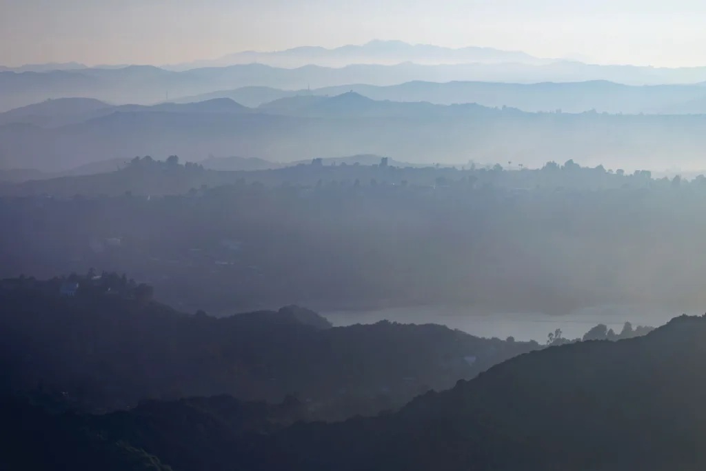 Photograph of layers of hills that fade in the distance with clouds of smoke obstructing the clear view of the sky and mountains.