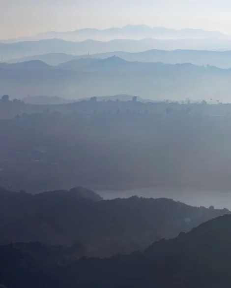 Photograph of layers of hills that fade in the distance with clouds of smoke obstructing the clear view of the sky and mountains.