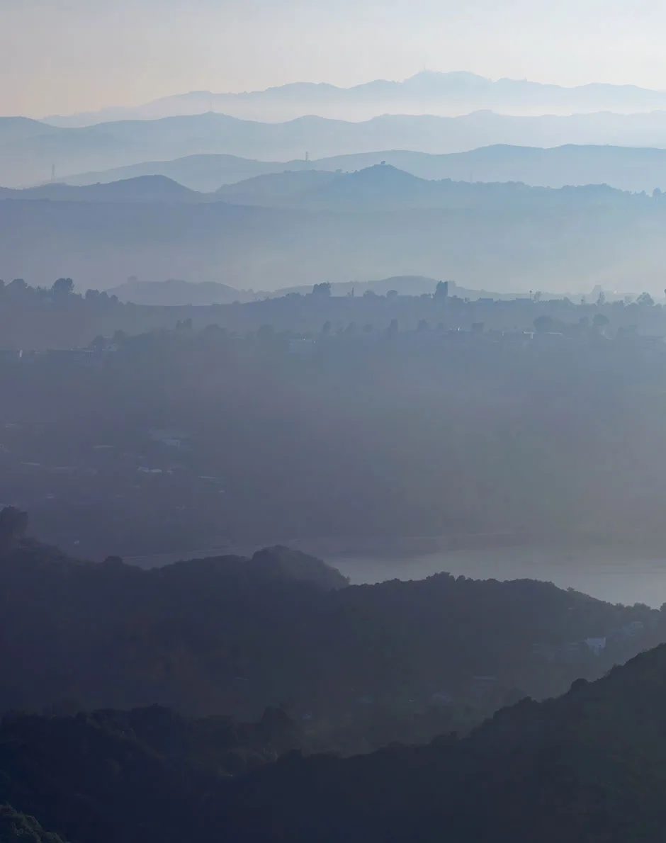 Photograph of layers of hills that fade in the distance with clouds of smoke obstructing the clear view of the sky and mountains.