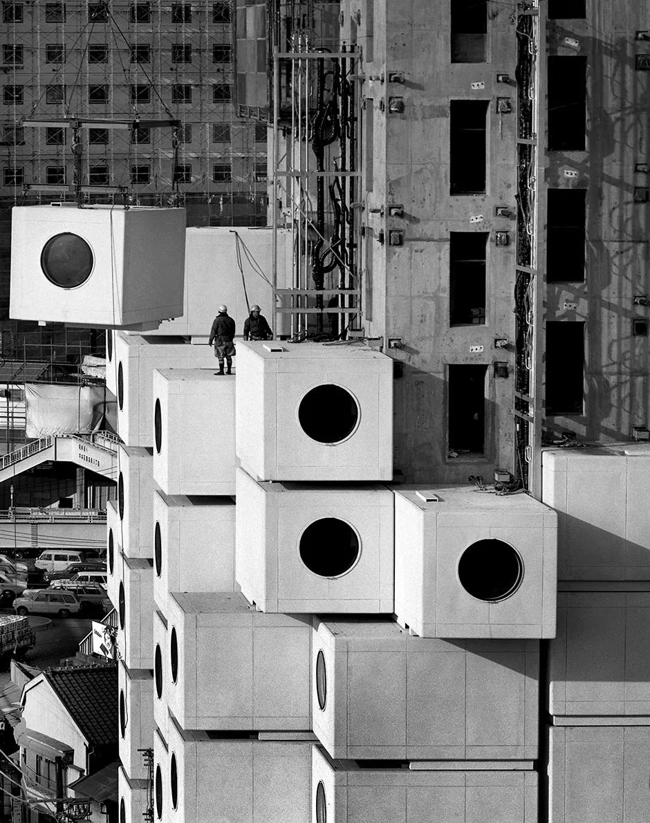 Black and white photograph of large concrete square structures with circle windows being lifted by crane and stacked on similar concrete blocks with two people in hard hats observing.