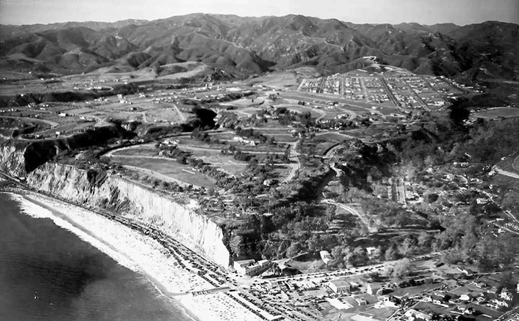 Black and white photograph of a neighborhood grid under development on a hill overlooking the ocean with mountains in the background.