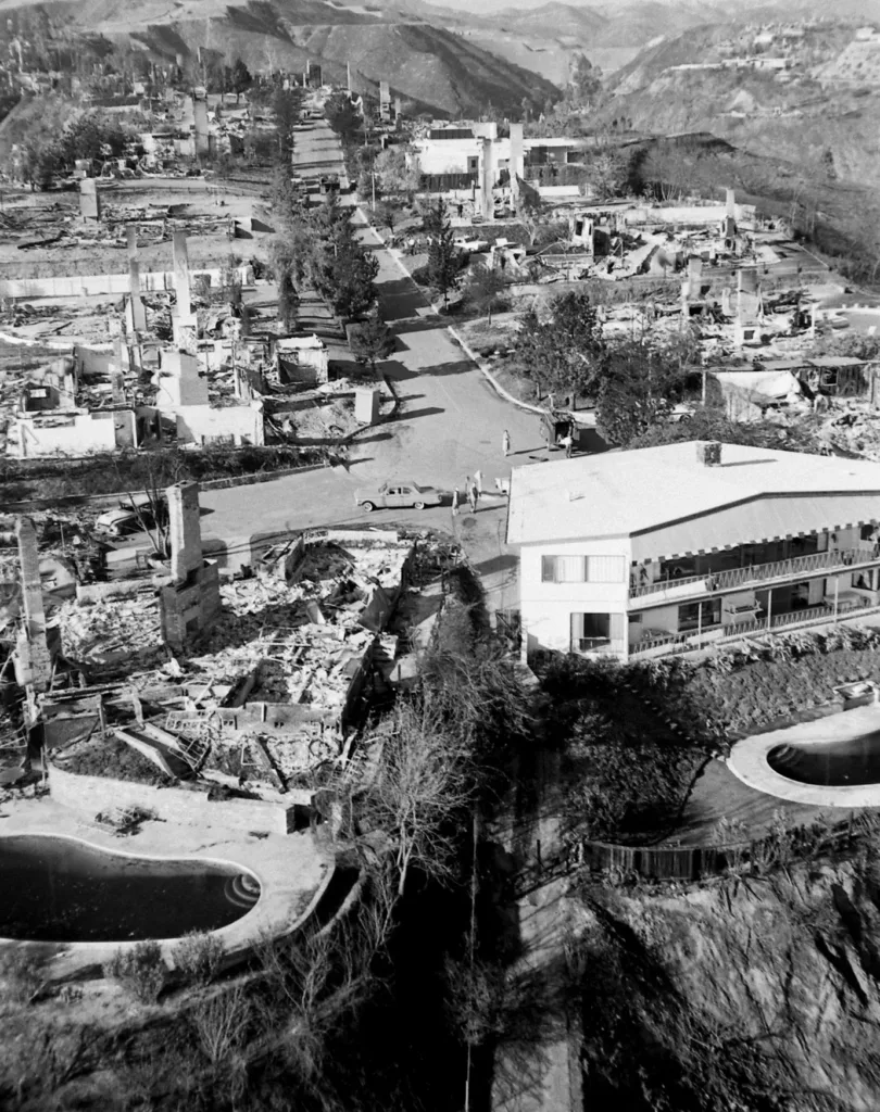 Black and white photograph of a neighborhood of burned buildings showing the charred remains of chimneys and swimming pools along a road with people in cars inspecting the damage.