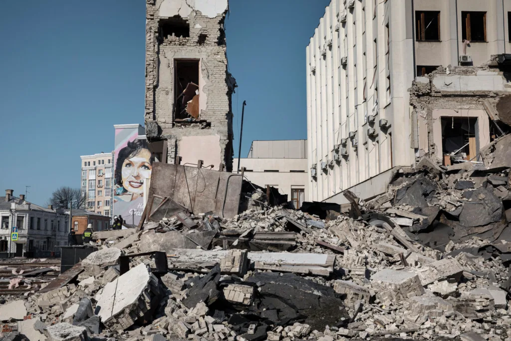 Rubble of a partially destroyed building with a mural of a woman painted onto a building in the background, under a clear blue sky.