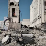 Rubble of a partially destroyed building with a mural of a woman painted onto a building in the background, under a clear blue sky.