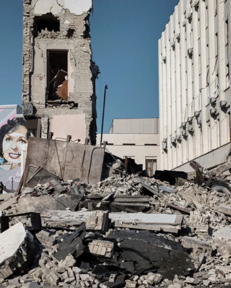 Rubble of a partially destroyed building with a mural of a woman painted onto a building in the background, under a clear blue sky.