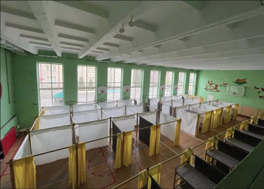 Large gym with green walls, converted into a temporary shelter with white cubicles. Yellow curtains provide privacy.