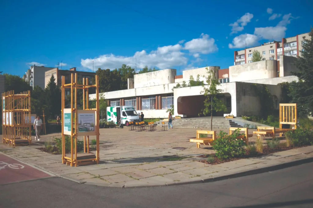 Public square with benches and wooden displays in front of a modern building under a blue sky with clouds, people walking, and a parked truck.