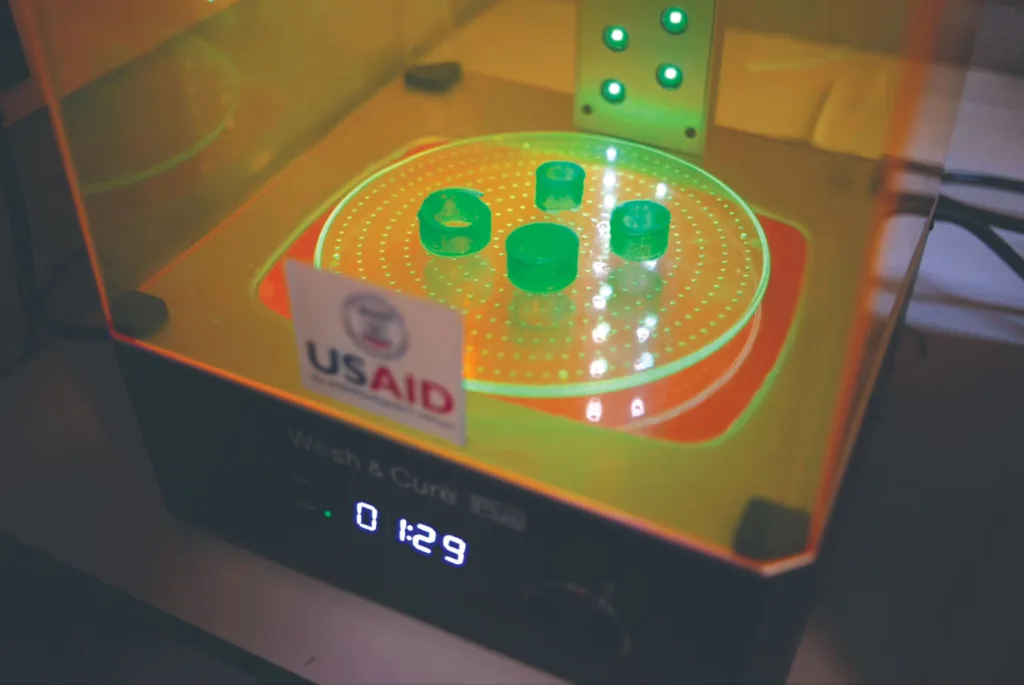 A 3-D printer that says USAID on the front glass panel with four green printed, green objects inside on a clear, rotating platform with yellow light glowing. A digital timer displays 01:29.
