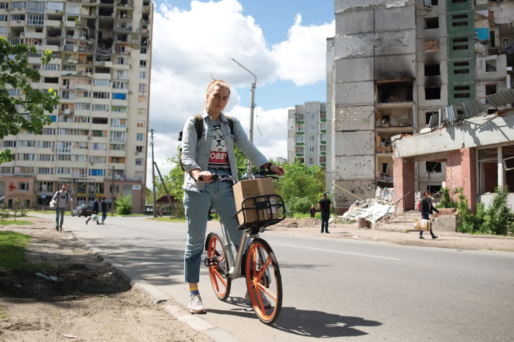 A person with a backpack pushes a bike with a basket carrying a cardboard box in front of damaged buildings under a blue sky with clouds.