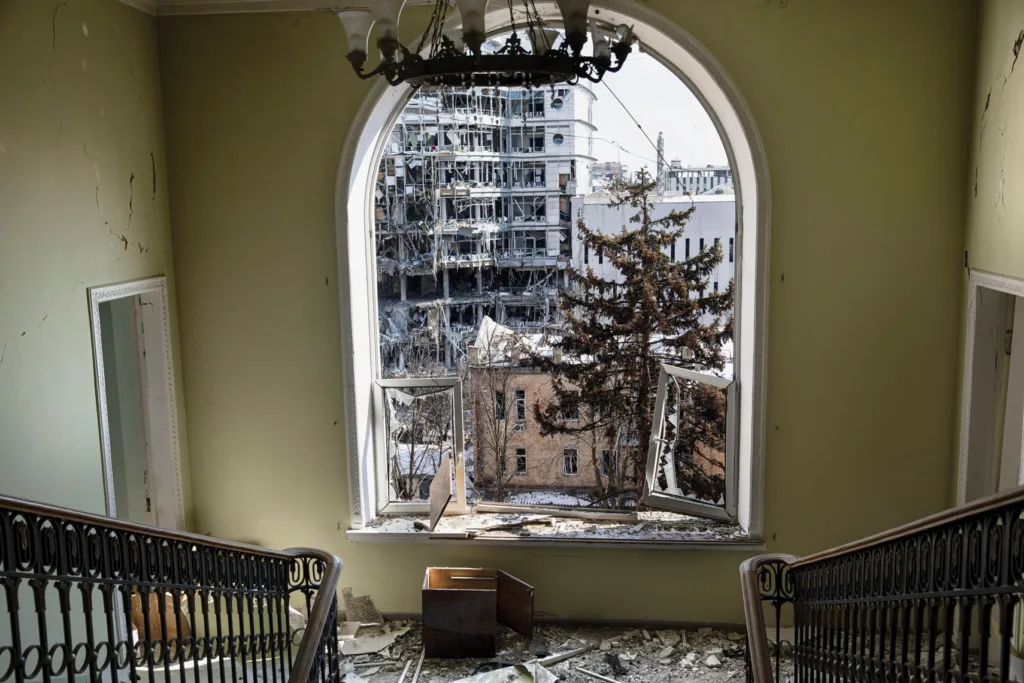 A staircase leads to a yellow wall with an arched window with broken glass, viewing a heavily damaged building and leafless trees outside.